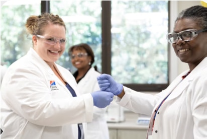 Zwei Wissenschaftler in Laborkitteln und Handschuhen lächeln und schütteln sich in einer Laborumgebung die Hände.