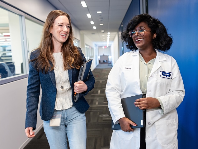 Deux femmes, l'une en tenue décontractée et l'autre en blouse blanche, marchent et discutent dans le couloir d'un bâtiment de Colgate-Palmolive, souriantes et un cahier à la main.